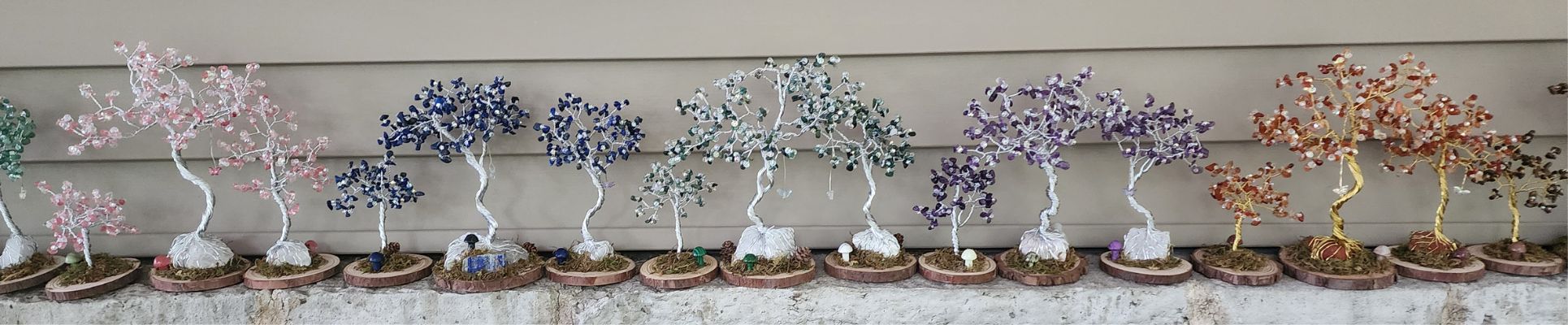 Row of wire and crystal trees. cheery quartz, lapis lazuli, moss agate, amethyst, carnelian and tiger eye lined on a whote brick ledge with a beige aluminum siding background